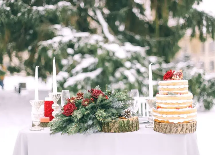 Kış bahçesinde bulunan masa üzerinde düğün pastası, mumlar, bardaklar ve çam ağacı dalları bulunuyor.